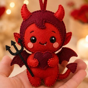 May include: A small, handcrafted felt devil ornament in shades of red and burgundy. The figure features two horns, pointed ears, small wings, a tail with a pointed tip, and a single white tooth. It holds a black felt trident and a dark red heart.