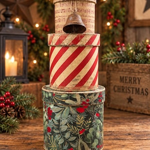 May include: A stack of three cylindrical boxes. The top box features musical notes and text, the middle box has red and white diagonal stripes, and the bottom box displays a dark green pattern of leaves and red berries. A small metal bell hangs from the top.