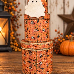 May include: A stack of three cylindrical orange boxes featuring a repeating pattern of black and red candies, lollipops, and candy corn. A white felt ghost cutout with two black eyes is attached to the front of the middle box in the stack.
