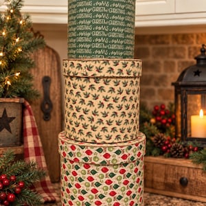 May include: A stack of three cylindrical cardboard gift boxes. The top box is dark green with white Merry Christmas text. The middle box features a pattern of green holly leaves and red berries. The bottom box displays red and green ornaments.