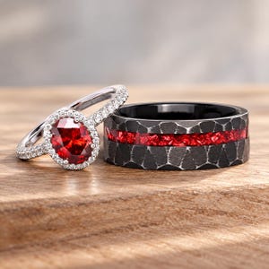 Puede incluir: Un anillo plateado con una piedra roja de corte ovalado rodeada de pequeñas piedras blancas, junto a una banda negra ancha con textura martillada y una incrustación central de material rojo triturado.