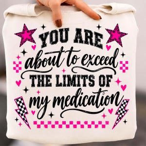 Może przedstawiać: Kremowa bluza z czarnym i jaskraworóżowym napisem "YOU ARE about to exceed THE LIMITS OF my medication". Wzór zawiera jaskraworóżowe gwiazdy w panterkę, czarno-białe błyskawice w szachownicę oraz małe serduszka rozmieszczone wokół tekstu.