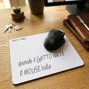Può includere: Un tappetino per mouse rettangolare bianco con il testo nero "[(QC Lab)]" nell'angolo in alto a sinistra e la frase "quando il GATTO non c'è il MOUSE balla" stampata al centro. Un mouse per computer nero poggia sul tappetino.