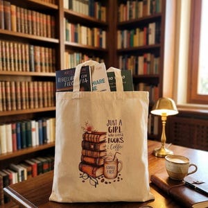 Może przedstawiać: Beżowa torba płócienna z nadrukiem stosu książek, kubka do kawy z napisem "COFFEE FIRST" oraz tekstem "JUST A GIRL who loves BOOKS & Coffee." Torba posiada dwa materiałowe uchwyty i jest wypełniona kilkoma książkami w twardej oprawie.