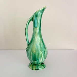 May include: A tall ceramic pitcher with a glossy finish featuring a marbled blend of emerald green, teal, and cream colors. The vessel has a bulbous base, a slender neck, a single curved handle, and an elongated, slanted spout opening at the top.