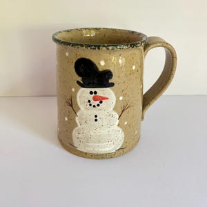 May include: A tan speckled ceramic mug featuring a hand-painted white snowman wearing a black top hat. The snowman has a bright orange carrot nose, black dot eyes, and a button mouth. The mug has a curved handle and a dark green rim.
