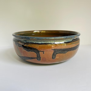 May include: A round ceramic bowl with a wide rim, featuring a glossy glaze in shades of warm terracotta, deep brown, and dark blue. The exterior displays a distinctive drip pattern where the dark glaze runs down the orange-brown base.