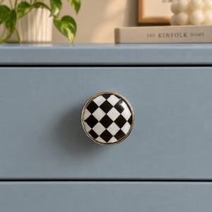 May include: A round cabinet knob featuring a black and white checkered pattern on its face, encased in a polished silver-toned metal rim. The knob is mounted centrally on a smooth, matte light blue drawer front, providing a distinct contrast.