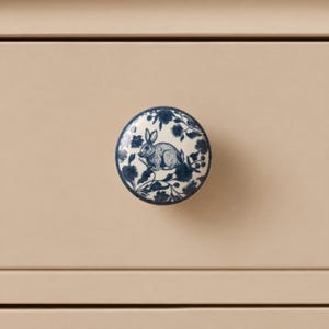 May include: A round ceramic drawer knob featuring a cream-colored base with a dark blue floral pattern and a central illustration of a rabbit. The knob is mounted on a flat, light beige surface, showcasing a smooth, glossy finish throughout.