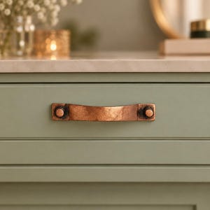 May include: A horizontal drawer pull handle with a weathered copper finish and a slightly curved shape, mounted onto a sage green cabinet drawer front. The handle features two circular attachment points on each end with a dark, textured patina.