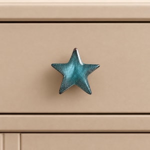 May include: A star-shaped drawer knob with a textured, metallic teal finish. The surface of the knob features a dark, mottled pattern that transitions into a vibrant blue-green hue, mounted on a flat, light beige, horizontal drawer front.