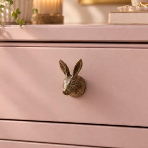 May include: A metallic, bronze-toned drawer knob shaped like a rabbit head with detailed ears and facial features, mounted on a smooth, matte light pink drawer front. The rabbit head is oriented forward, showing textured fur details.