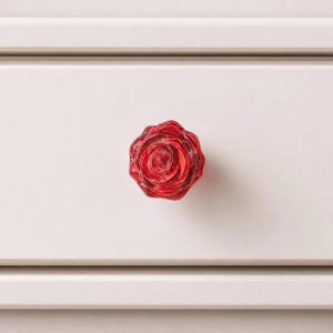 May include: A translucent red glass drawer knob shaped like a blooming rose. The knob features intricate, layered petal details that create a realistic floral appearance. It is mounted on a flat, light-colored surface with horizontal paneling.