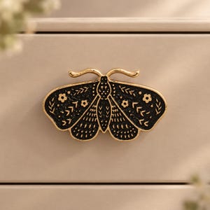 May include: A moth-shaped drawer pull featuring a black textured surface with gold-colored outlines and detailing. The design includes engraved floral and leaf patterns across the wings, with two curved antennae extending from the top of the head.