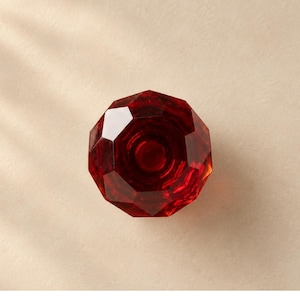 May include: A faceted, circular red glass knob with a multi-sided geometric design. The surface features a polished, reflective finish with deep crimson tones and a central circular indentation, creating a clear, jewel-like appearance.