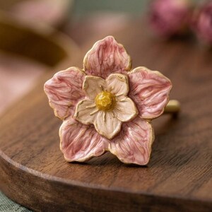 May include: A decorative cabinet knob shaped like a five-petaled flower. The outer petals are a dusty rose pink with a textured, veined finish, while the inner petals are a lighter cream color. A small, textured golden yellow center completes the piece.