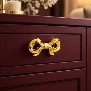 May include: A shiny gold-toned metal drawer pull shaped like a decorative bow, mounted on a dark burgundy cabinet drawer. The bow features smooth, reflective surfaces and a central knot detail, contrasting against the matte finish of the furniture.