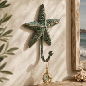 May include: A wall-mounted hook shaped like a five-pointed starfish with a textured, weathered teal and bronze finish. The hook extends downward from the center of the starfish, featuring a rounded tip that holds a gold-toned pendant necklace.