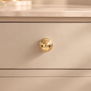 May include: A polished gold-toned spherical cabinet knob centered on a smooth, matte beige drawer front. The knob features a reflective, rounded surface that catches light, contrasting with the flat, neutral-colored finish of the drawer panel.
