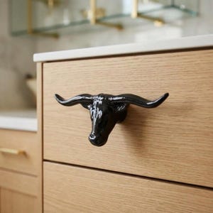 May include: A glossy black cabinet knob shaped like a longhorn bull head with prominent curved horns. The knob is mounted on a light-colored wood drawer front with a visible grain pattern, serving as a functional handle for the furniture piece.