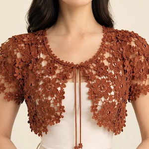 May include: A rust-colored crochet bolero featuring an intricate floral pattern throughout the garment. This short-sleeved shrug has a rounded neckline and includes two thin matching strings at the center front that can be tied together.