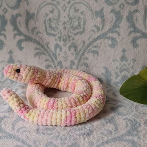 May include: A coiled plush snake toy crafted from soft, variegated yarn in shades of pastel pink, cream, and pale yellow. The stuffed animal features a rounded head with small, dark, circular plastic eyes and a segmented, textured body.