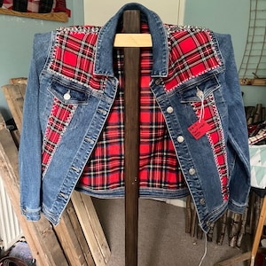 Peut inclure: Une veste en jean bleu avec des empiècements en tissu écossais rouge et noir sur le devant et les épaules, agrémentée de points de croix blancs et de clous métalliques argentés le long des coutures. Une étiquette rouge avec du texte est attachée.