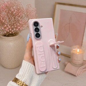 May include: A light pink smartphone case featuring a vertical camera cutout with three lenses and an integrated, extendable kickstand on the back. The word FOLD is embossed vertically in capital letters on the kickstand mechanism.