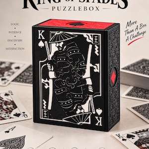 May include: A black and red rectangular puzzle box titled King of Spades Puzzlebox. The front features a white graphic of a samurai figure. Text includes Logic, Patience, Discovery, Satisfaction, and More Than A Box A Challenge. Dimensions are 15 x 12 x 5 cm.
