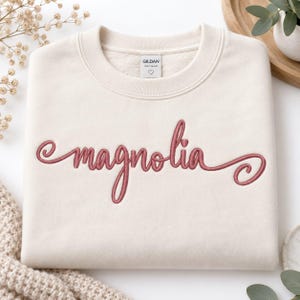 Peut inclure: Un sweat-shirt à col rond de couleur crème avec le mot magnolia brodé en écriture cursive rose poudré sur la poitrine. Le vêtement présente une étiquette de marque Gildan à l'intérieur du col avec une petite icône de cœur sous le texte.