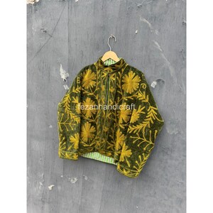 Puede incluir: Una chaqueta de color verde oliva oscuro con un vibrante patrón de bordado floral amarillo en la parte delantera y las mangas. La chaqueta tiene cuello alto, mangas largas y un forro estampado en verde claro visible en el dobladillo, colgada.