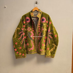 Puede incluir: Una chaqueta de terciopelo verde oliva con bordados florales y abstractos en tonos rosa, amarillo y azul oscuro. La chaqueta tiene cuello alto, mangas largas y un forro a rayas blancas y negras visible a lo largo de la abertura frontal.