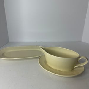 May include: A cream-colored gravy boat with a matching oval saucer and a separate elongated oval serving platter. The set features a smooth, solid-colored finish with a curved handle on the gravy boat and a wide, shallow rim on the platter.