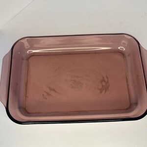 May include: A rectangular Pyrex baking dish made of translucent purple glass. The dish features rounded corners and integrated handles on the short sides. The center of the base is embossed with the Pyrex brand name and manufacturing details.