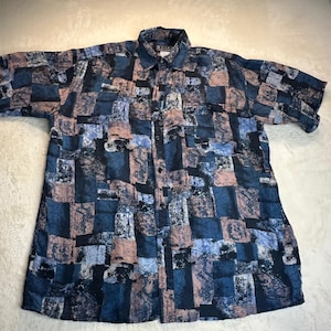 May include: A short-sleeve button-down shirt featuring a patchwork-style pattern in shades of navy blue, light blue, and muted mauve. The design consists of rectangular blocks with varied textures and abstract line details throughout the fabric.