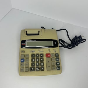 Peut inclure: Une calculatrice imprimante Sharp EL-2192G beige avec un cordon d'alimentation noir. L'appareil dispose d'un écran numérique, d'un compartiment pour rouleau de papier et d'un pavé numérique avec des touches grises et bleues, dont une touche égale rouge.