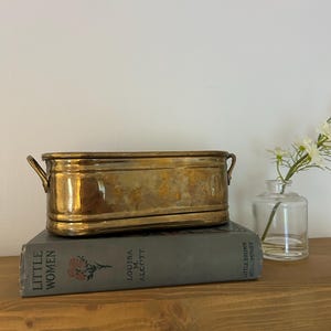 May include: An oval brass planter with two small side handles, resting on a gray hardcover book titled Little Women by Louisa M. Alcott, published by Little Brown and Company, accompanied by a small clear glass vase with white flowers.