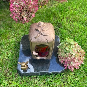 Op de afbeelding: Een bronskleurige metalen graflantaarn met een roosreliëf op de bovenkant en een glazen inzetstuk, geplaatst op een rechthoekige zwarte granieten voet naast een klein bronskleurig engelfiguurtje, tegen een achtergrond van groen gras.