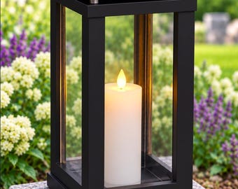 May include: A black rectangular lantern with a solid top and clear glass panels on four sides. Inside the lantern, a single white pillar candle with a warm, glowing flame is positioned centrally on the base of the structure.