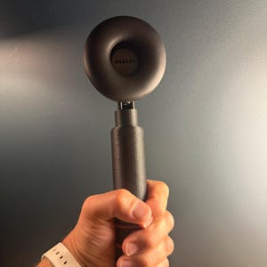 May include: A black, circular, padded headphone earcup attached to a cylindrical, textured black handle. The centre of the earcup features the text AIAIAI printed in a dark font. The device is held vertically, showcasing its modular design.