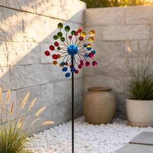 May include: A kinetic garden spinner featuring a central blue sphere with radiating metal arms, each tipped with small, multicoloured spheres in shades of red, yellow, green, and blue, mounted on a tall, slender black metal pole.