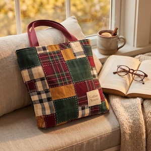 Può includere: Una borsa tote trapuntata con un design patchwork di quadrati di tessuto a quadri e tinta unita nelle tonalità verde scuro, bordeaux, giallo senape e crema. La borsa ha due manici bordeaux e una piccola etichetta rettangolare con scritto Gather Round.