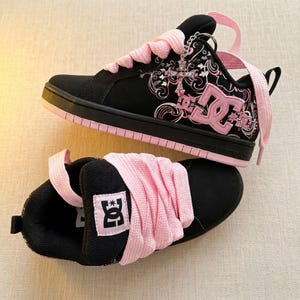 Puede incluir: Un par de zapatillas DC negras y rosas con cordones anchos de color rosa, un logotipo DC rosa en el lateral y un dije de cruz plateada unido a los ojales. Los zapatos tienen una base negra con patrones florales rosas y una suela de goma rosa.
