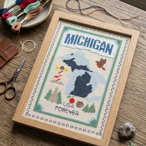 May include: A framed cross-stitch embroidery depicting a map of Michigan with the text MICHIGAN and USA FOREVER. The design includes a lighthouse, trees, a bird, and fruit icons, surrounded by a geometric border in a light wood frame.