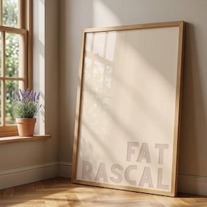 May include: A rectangular framed print featuring the text FAT RASCAL in large, bold, light-colored capital letters. The artwork is encased in a simple, light-toned wooden frame, resting on a wooden floor with a herringbone pattern.