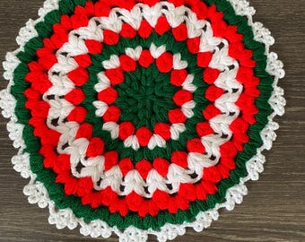 May include: A round, hand-crocheted doily featuring a concentric circular pattern. The design consists of alternating rings of vibrant red, forest green, and bright white yarn, finished with a delicate, scalloped white lace-like border edge.