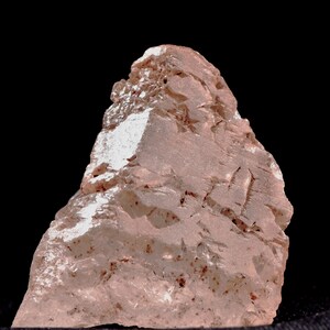 Può includere: Un campione di minerale rosa chiaro, tagliato in modo grezzo, su sfondo nero. La pietra presenta una struttura cristallina con fratture visibili e una superficie leggermente riflettente. La forma generale è irregolare, con aspetto strutturato.