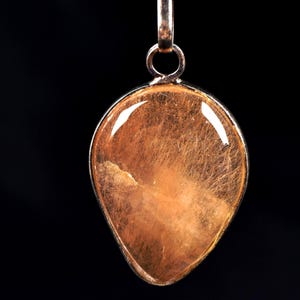 Può includere: Un ciondolo a forma di goccia con una montatura in metallo color argento. Il ciondolo presenta una pietra traslucida color ambra con delicate inclusioni a forma di capelli. Lo sfondo è nero pieno, evidenziando i dettagli del ciondolo.