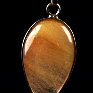 Può includere: Un ciondolo a forma di goccia con una pietra traslucida color marrone dorato che presenta inclusioni interne simili ad aghi. La pietra è incastonata in una montatura in metallo color argento con un anellino circolare sulla parte superiore.