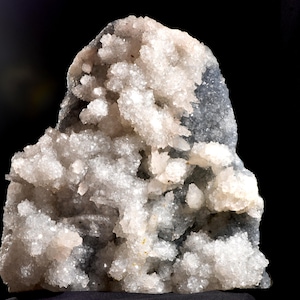 May include: A raw mineral specimen featuring a cluster of translucent, white, crystalline formations protruding from a dark, solid gray rock base. The jagged, sparkling crystal points vary in size and density, creating a textured, uneven surface.